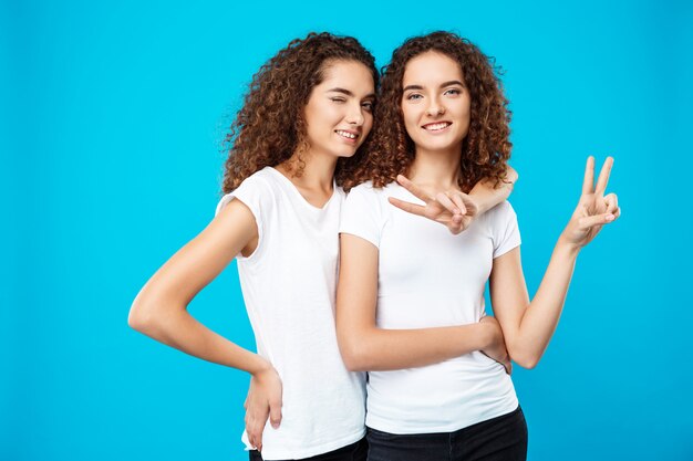 Duas meninas bonitas gêmeas sorrindo, mostrando paz sobre parede azul