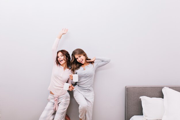 Duas meninas de pijama com xícaras na parede cinza. Eles se espreguiçando e sorrindo.