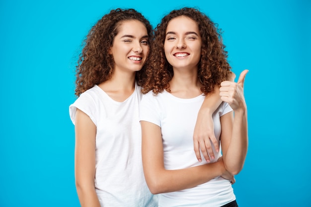 Duas meninas gêmeas sorrindo, piscando, mostrando como parede azul
