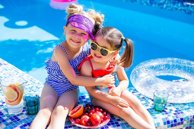 Duas meninas sentadas à beira da piscina no verão usando óculos escuros e bebendo limonada