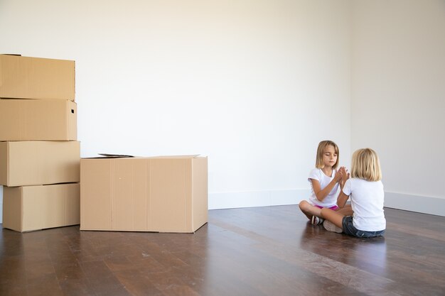 Duas meninas sentadas no chão perto de uma pilha de caixas em seu novo apartamento e brincando juntas