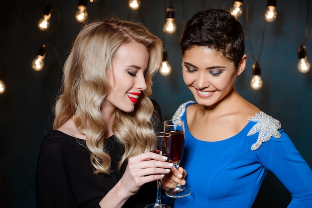 Duas mulheres bonitas em vestidos de noite, sorrindo, segurando copos de vinho