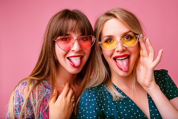 Duas mulheres bonitas engraçadas se divertindo fazendo selfie e mostrando línguas compridas, usando óculos escuros e vestidos coloridos, clima de primavera verão, parede rosa.