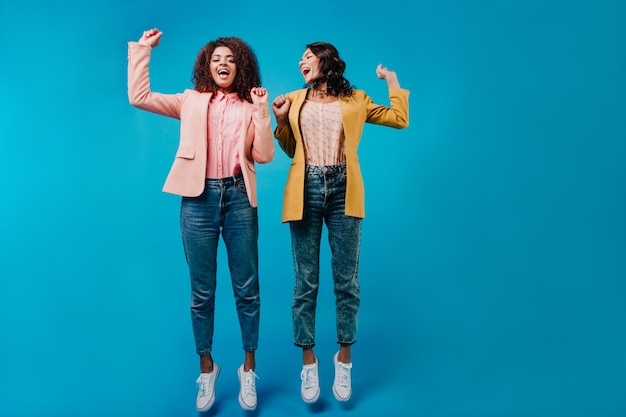 Duas mulheres de jeans pulando na parede azul