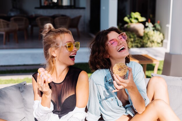 Duas mulheres felizes em forma de óculos de sol rosa e amarelo sorrindo, se divertindo, rindo com donuts, ao ar livre