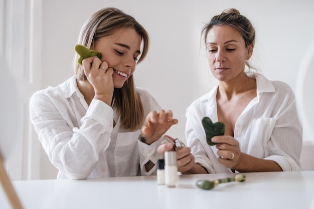 Duas mulheres loiras caucasianas de diferentes idades estão provando produtos cosméticos enquanto estão sentadas no fundo branco. Cuidados com a pele e conceito de hidratação