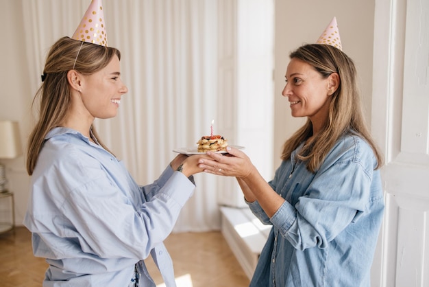 Duas mulheres loiras de pele clara se olham e seguram um pedaço de bolo de aniversário com vela dentro de casa. feriados, celebração e conceito de mulheres.