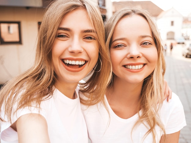 Duas mulheres loiras hipster sorridente jovens em roupas de camiseta branca de verão. Meninas tirando fotos de auto-retrato de selfie no smartphone.