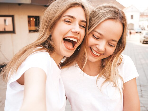 Duas mulheres loiras hipster sorridente jovens em roupas de camiseta branca de verão. Meninas tirando fotos de auto-retrato de selfie no smartphone.