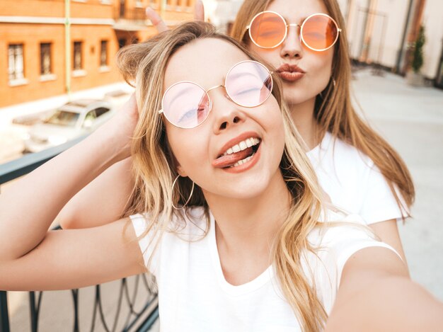 Duas mulheres loiras hipster sorridente jovens em roupas de camiseta branca de verão. Meninas tirando fotos de auto-retrato de selfie no smartphone.