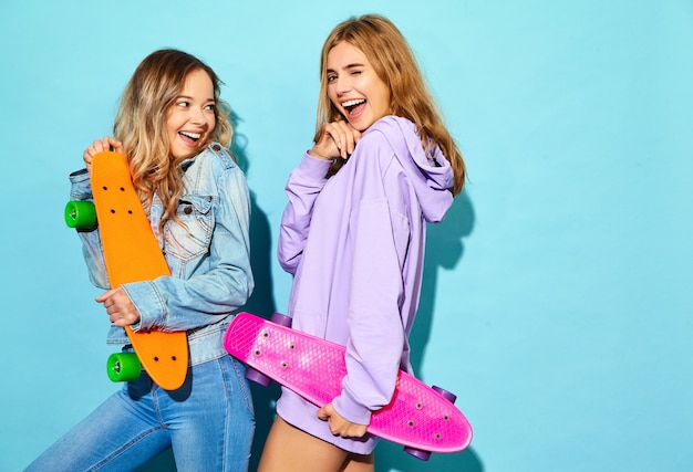 Duas mulheres loiras sorridentes elegantes jovens com skates centavo. Modelos em roupas de esporte hipster de verão posando perto de parede azul. Fêmea positiva
