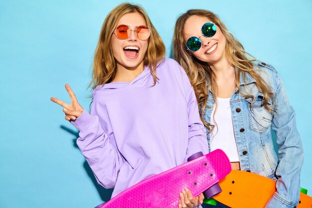 Duas mulheres loiras sorridentes elegantes jovens com skates centavo. Modelos em roupas de esporte hipster de verão posando perto de parede azul. Mulheres positivas enlouquecendo