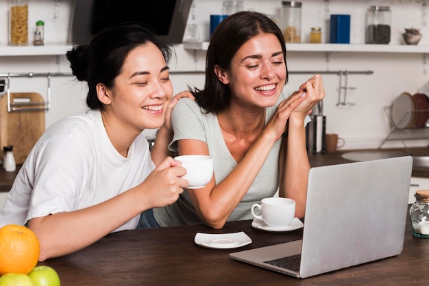 Duas mulheres na cozinha de casa olhando para o laptop enquanto tomam café