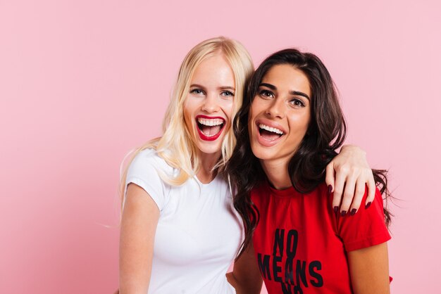 Duas mulheres rindo, abraçando e olhando para a câmera sobre rosa