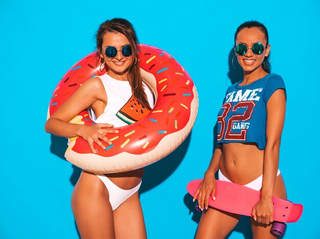 Duas mulheres sexy sorridentes bonitas em cuecas e tópico de verão. Garotas de óculos escuros. Modelos positivos se divertindo com skates centavo coloridos. Com colchão inflável donut lilo