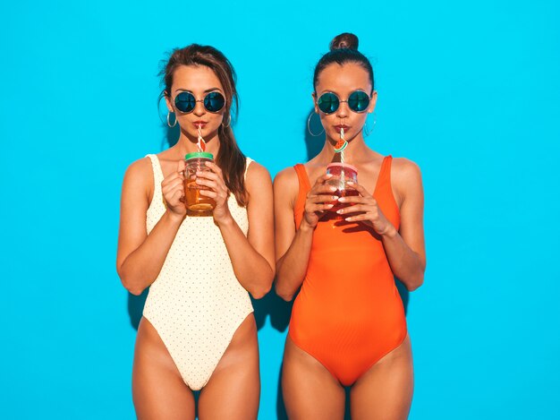 Duas mulheres sorridentes sexy bonitas em trajes de banho swimwear colorido de verão. Garotas da moda em óculos de sol. Ficando louco. Modelos engraçados isolados. Beber coquetel fresco bebida suave