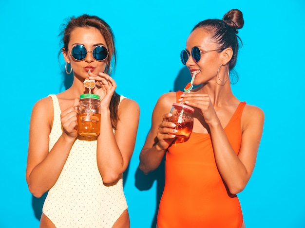 Duas mulheres sorridentes sexy bonitas em trajes de banho swimwear colorido de verão. Garotas da moda em óculos de sol. Ficando louco. Modelos engraçados isolados. Beber coquetel fresco bebida suave