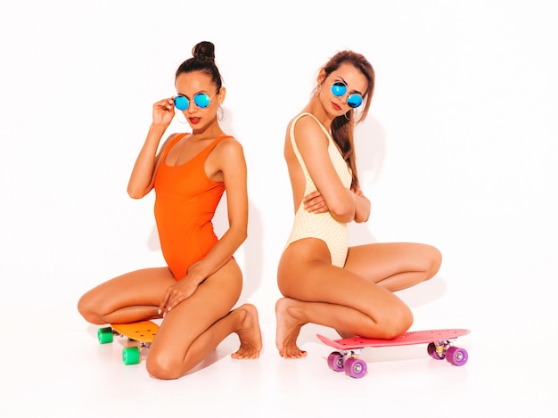 Duas mulheres sorridentes sexy bonitas em trajes de banho swimwear colorido de verão. meninas da moda em óculos de sol. modelos positivos, sentado no chão com skates centavo coloridos. isolado