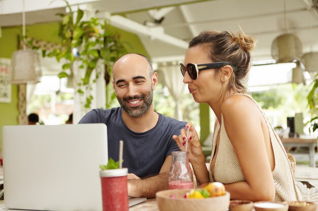 Duas pessoas assistindo vídeo online ou vendo fotos na Internet, usando wi-fi no laptop durante o almoço. Feliz homem barbudo e mulher elegante em tons relaxantes no café com o notebook.