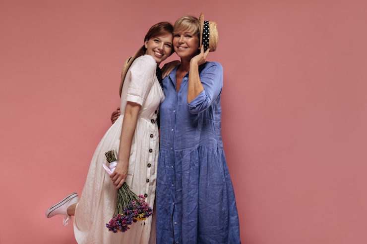 Foto grátis duas senhoras modernas com penteado curto legal em vestidos da moda, sorrindo, abraçando e segurando o buquê e o chapéu de palha no pano de fundo isolado.