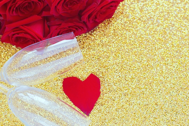 Duas taças de champanhe, um buquê vermelho de rosas e um coração de pétalas repousa sobre um ouro brilhante ...