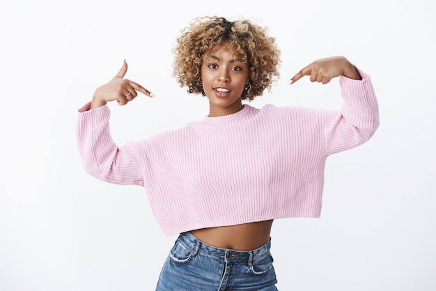 Foto grátis e quanto a mim. retrato de mulher bonita, elegante e confiante, com cabelo loiro afro, parecendo ousada e segura de si mesma, apontando para si mesma posando orgulhosamente sobre uma parede branca