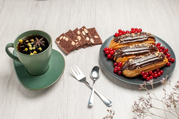 Éclairs de chocolate e groselha no prato cinza biscoitos de amendoim uma xícara de chá, colher e um garfo em vetor diagonal na mesa de madeira branca.