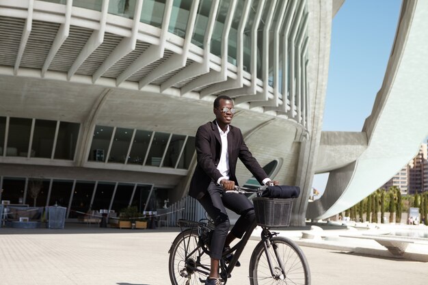 Ecologicamente consciente, jovem empresário europeu preto vestindo terno preto e óculos de sol, andar de bicicleta para o café na bicicleta retrô durante a pausa para o almoço em dia ensolarado