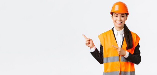 Edifício, construção e conceito industrial. Arquiteta asiática sorridente com capacete de segurança, roupa reflexiva apontando o dedo no canto superior esquerdo, mostrando o projeto no local de trabalho, fundo branco