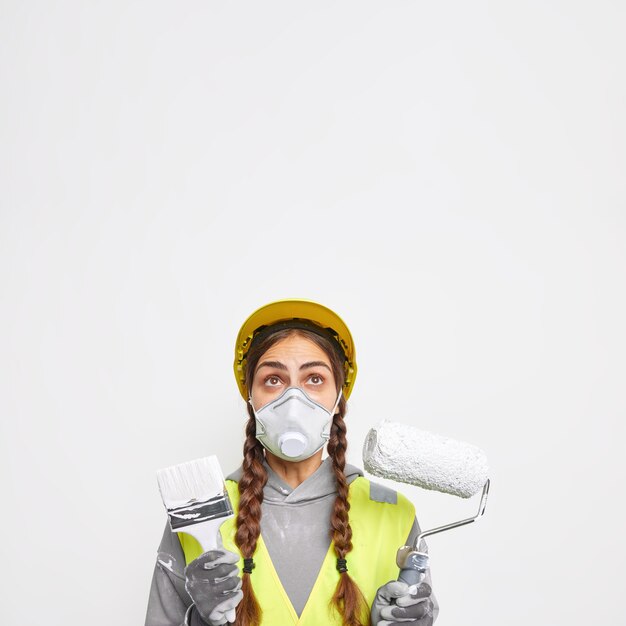 Foto grátis edifício e conceito de construção. mulher surpreendida com duas marias-chiquinhas usa máscara protetora e capacete vestido em poses de uniforme com o equipamento de construção focado acima, isolado sobre a parede branca