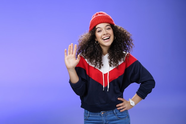 Foto grátis ei, prazer em conhecê-la encantadora sofá de esqui feminino em um gorro vermelho bonito com cabelo encaracolado acenando com ...