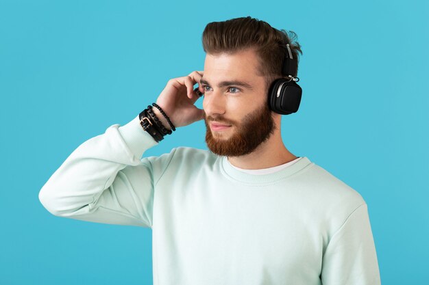 elegante atraente barbudo jovem ouvindo música em fones de ouvido sem fio estilo moderno clima confiante isolado na parede azul