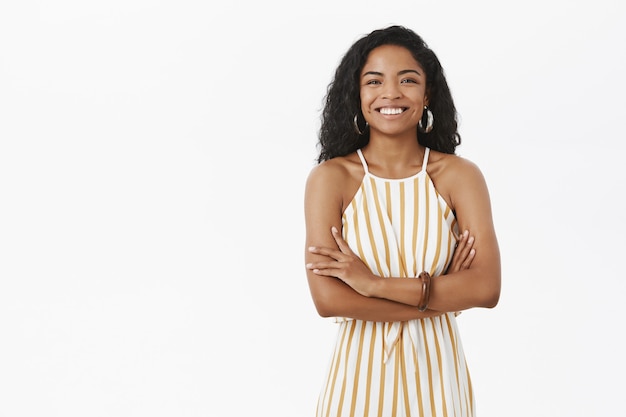 Elegante bem-sucedida encantadora empresária afro-americana com penteado encaracolado em macacão listrado amarelo, cruzando os braços no peito