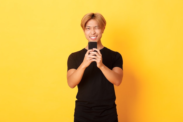 Elegante cara bonito asiático tirando foto no smartphone e sorrindo, fotografando com o celular, em pé na parede amarela