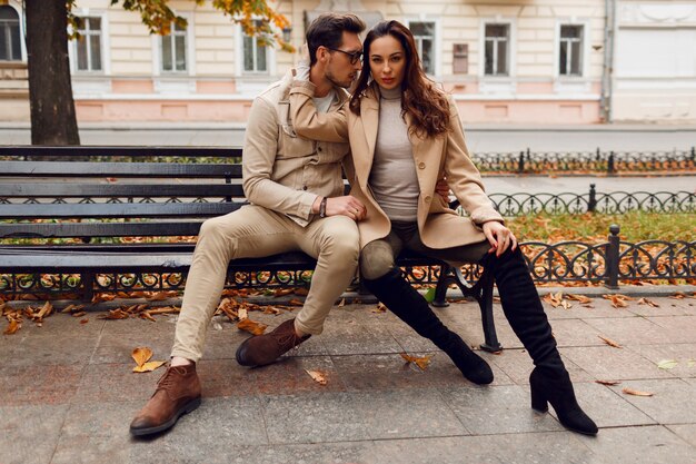 Elegante casal apaixonado posando ao ar livre. Tendências da moda outono. Modelo Moreno com cara elegante casaco bege, sentado no banco.
