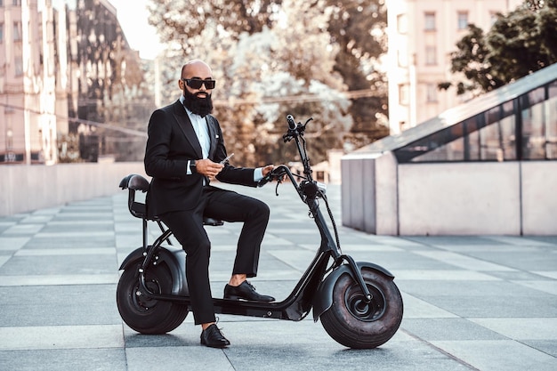 Elegante empresário careca em óculos de sol está fumando vaporizador enquanto está sentado em sua bicicleta elétrica.