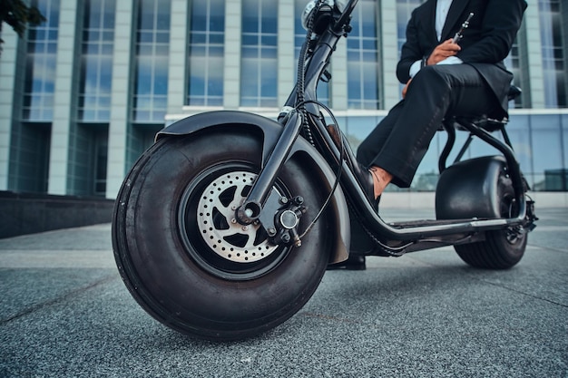 Elegante empresário de terno preto está sentado em sua scooter elétrica perto de seu escritório.