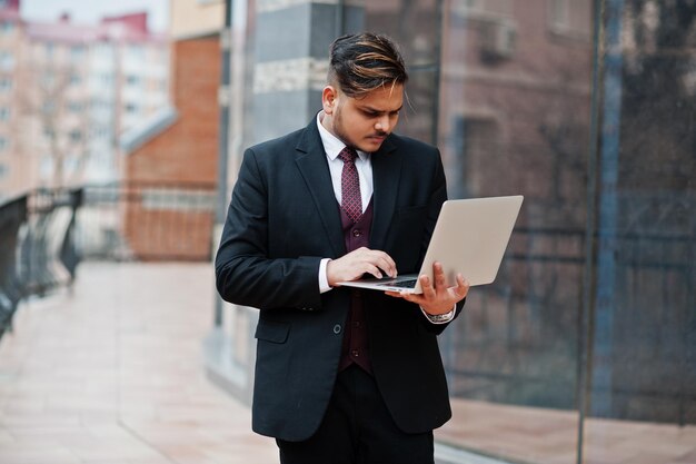 Elegante empresário indiano com roupa formal com laptop nas mãos de pé contra janelas no centro de negócios