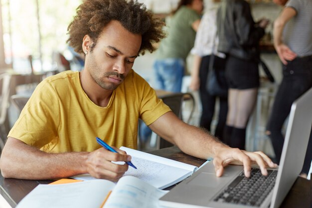 elegante estudante masculino sentado no restaurante na mesa de madeira, escrevendo algo em seu livro de cópia, digitando no teclado de seu laptop, olhando seriamente para baixo. Preparação para exames ou aulas