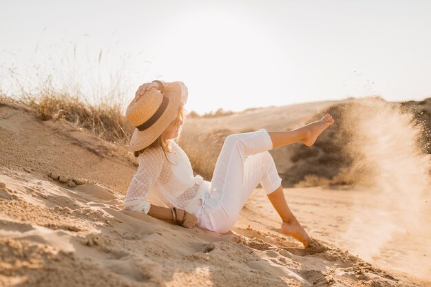 Elegante feliz atraente sorridente mulher posando no deserto vestida com roupas brancas, chapéu de palha e óculos escuros no pôr do sol