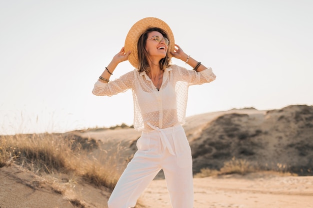 Elegante feliz linda mulher sorridente posando na areia do deserto com roupa branca, chapéu de palha e óculos escuros no pôr do sol