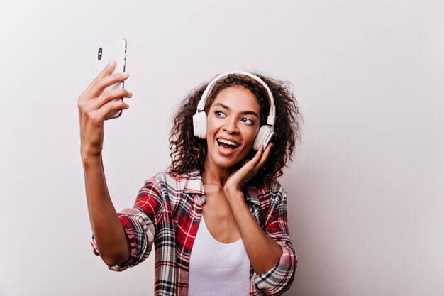 Elegante garota negra ouvindo música enquanto tira uma foto de si mesma. Mulher entusiasmada usando telefone para selfie.