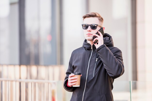 Elegante, homem jovem, com, óculos de sol, usando, telefone móvel, segurando, takeaway, xícara café