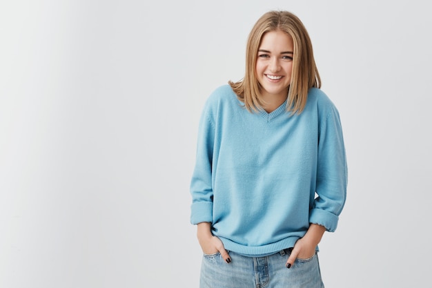 Elegante jovem aparência feminina de aparência Europeia vestindo jeans e blusa azul, sorrindo amplamente, demonstrando seus dentes brancos perfeitos, segurando as mãos nos bolsos. Juventude e felicidade