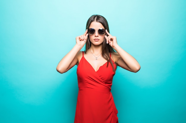 Elegante jovem atraente com vestido vermelho verão com as mãos nos óculos de sol isolados sobre a parede azul pastel.