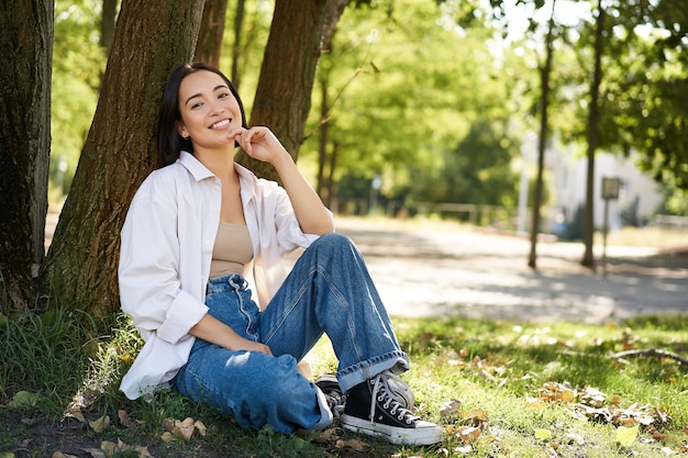 Elegante jovem estudante universitária senta-se no parque no gramado se inclina na árvore e sorri descansando ao ar livre