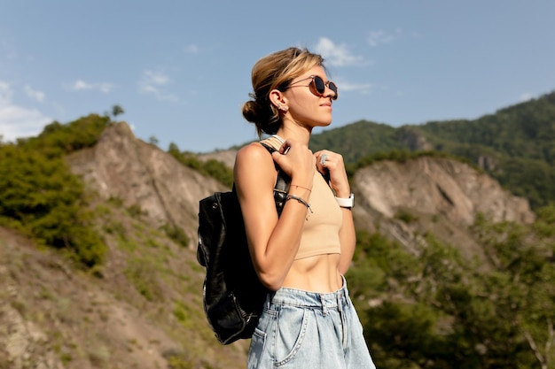 Elegante jovem europeia em óculos de sol vestindo camiseta e shorts com mochila está viajando entre a natureza selvagem no dia ensolarado de verão