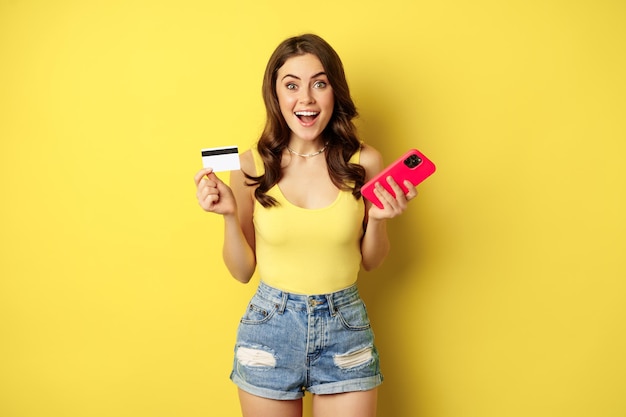 Elegante jovem morena, segurando o cartão de crédito e smartphone, pagando, comprando ou encomendando algo na loja, de pé sobre fundo amarelo.