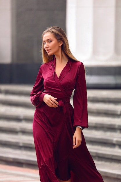Elegante jovem num vestido longo cor de vinho com mangas e cós. Segurando a mão sob os seios. Penteado loiro e maquiagem nude natural. Descendo a rua perto do prédio.