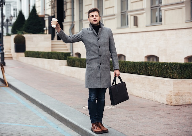 Elegante jovem segurando café para viagem em copo de papel e pegando táxi na rua vazia